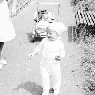 Baby in a suit walks near a woman and bench.
