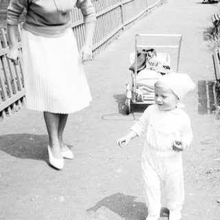 Baby in a suit walks near a woman and bench.