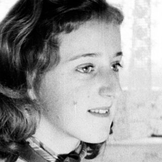 Here's a brief summary of the image: Young woman sits, wearing a sweater and patterned skirt.