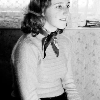 Here's a brief summary of the image: Young woman sits, wearing a sweater and patterned skirt.