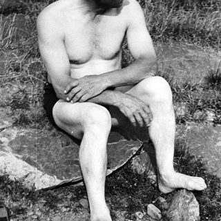 Here's a brief summary of the image: Bare-chested man sits on a rock, barefoot in nature.