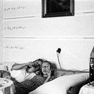 Here's a brief summary of the image: Two people relax on a bed, talking on a vintage telephone.