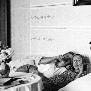 Here's a brief summary of the image: Two people relax on a bed, talking on a vintage telephone.