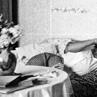 Here's a brief summary of the image: Two people relax on a bed, talking on a vintage telephone.