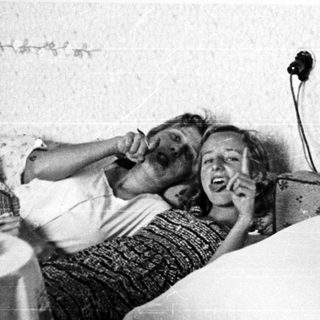Here's a brief summary of the image: Two people relax on a bed, talking on a vintage telephone.