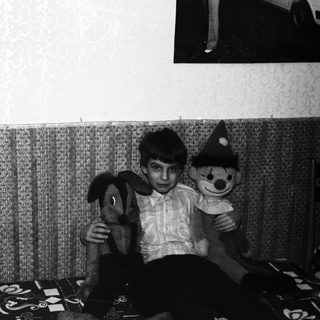 Boy sits with stuffed animals in a sparsely furnished room.
