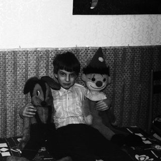 Boy sits with stuffed animals in a sparsely furnished room.