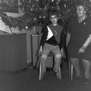 Here's a brief summary of the image: Boy sits near a decorated tree; woman stands nearby.