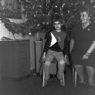 Here's a brief summary of the image: Boy sits near a decorated tree; woman stands nearby.