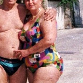 Couple in swimwear stands near seaside buildings and mountains.