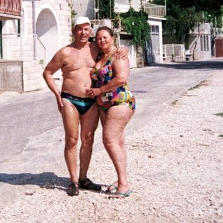 Couple in swimwear stands near seaside buildings and mountains.