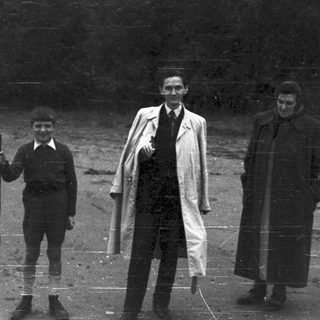 Three formally dressed figures stand outdoors in a wooded area.