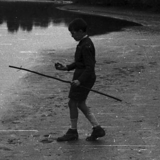 Boy walks along a riverbank holding a stick.