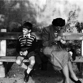 Boy sits on a bench while a woman knits.