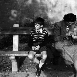 Boy sits on a bench while a woman knits.