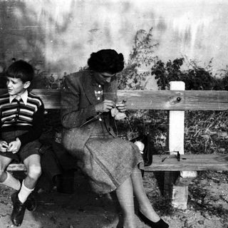 Boy sits on a bench while a woman knits.