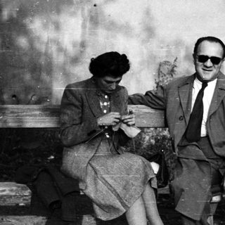 Couple seated on a bench, woman knitting, man reading.