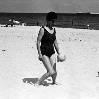 Woman walks on a beach, holding a ball.