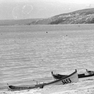 Children stand near rowboats on a beach with cliffs.