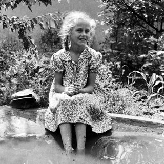 Girl sitting in water, smiling, surrounded by foliage.