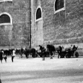 Here's a brief summary of the image: Horse-drawn cart and people near a stone building.