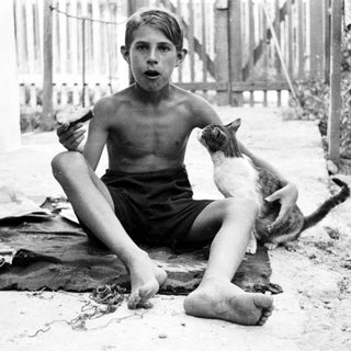 Here's a brief summary of the image: Bare-chested boy sits with a cat outdoors.