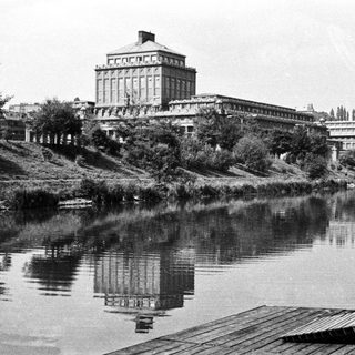 Large building reflected in water, with vegetation in foreground.