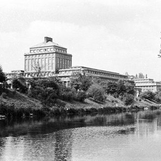 Grand building overlooks a river, viewed from across the water.