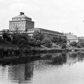 Large building overlooking a river with grassy banks.