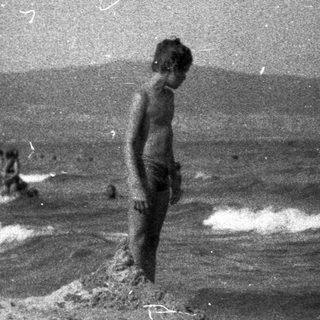Boy stands on a beach, facing the ocean.