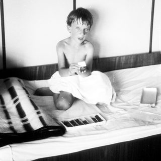 Here's a brief summary of the image: Black and white photo of a young boy seated on a bed.