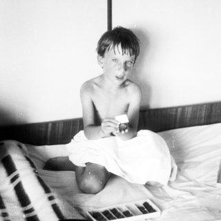 Here's a brief summary of the image: Black and white photo of a young boy seated on a bed.