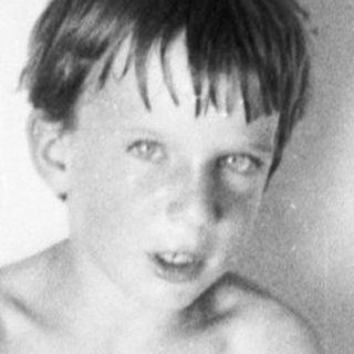 Here's a brief summary of the image: Black and white photo of a young boy seated on a bed.