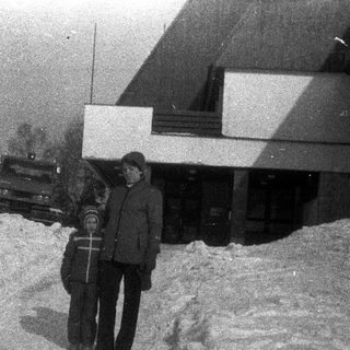 Two people stand in deep snow near a building.