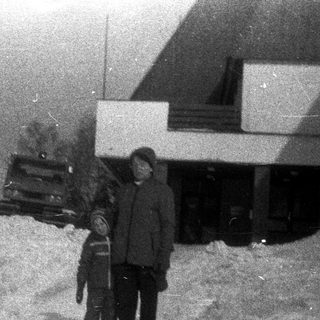 Two figures stand in a snowy landscape near a building.