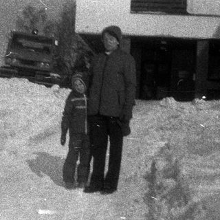 Two figures stand in a snowy landscape near a building.