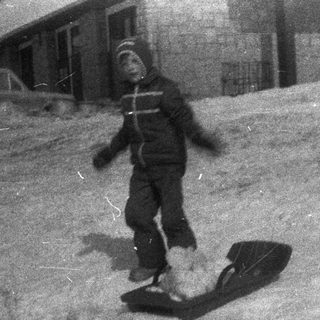 Child sledding down a snowy hill with a building nearby.