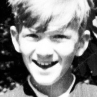 Boy in vintage clothing stands outdoors, smiling at the camera.