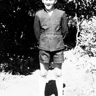 Boy in vintage clothing stands outdoors, smiling at the camera.