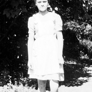 Here's a brief summary of the image: Young girl stands outdoors in a simple dress and shoes.
