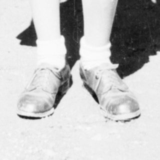 Here's a brief summary of the image: Young girl stands outdoors in a simple dress and shoes.