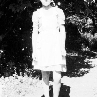 Here's a brief summary of the image: Young girl stands outdoors in a simple dress and shoes.