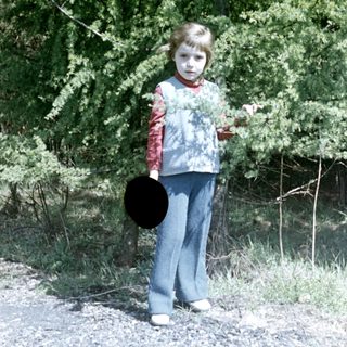 Boy standing outdoors, holding a hat, wearing a vest.