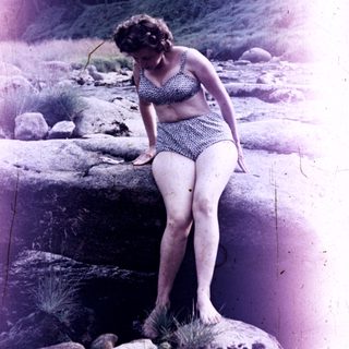 Woman in swimwear stands on rocks near a stream.