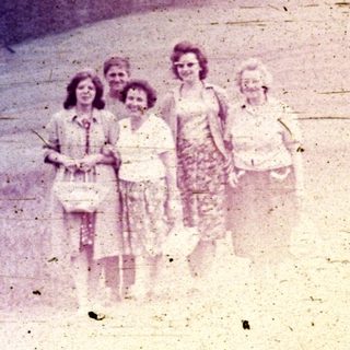 Four young women stand in a field with a forested hillside.