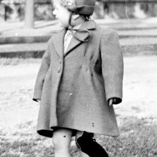 Child in coat stands on grass, looking away.