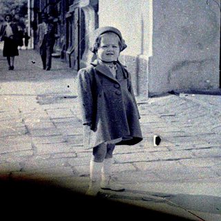 Child stands outdoors near building, people walk in background.