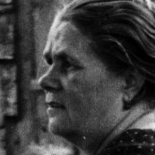 Woman seated near a weathered building and foliage.