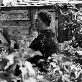 Woman seated near a weathered building and foliage.