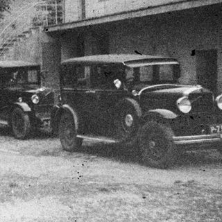 Group of people standing near a vintage car, building.
